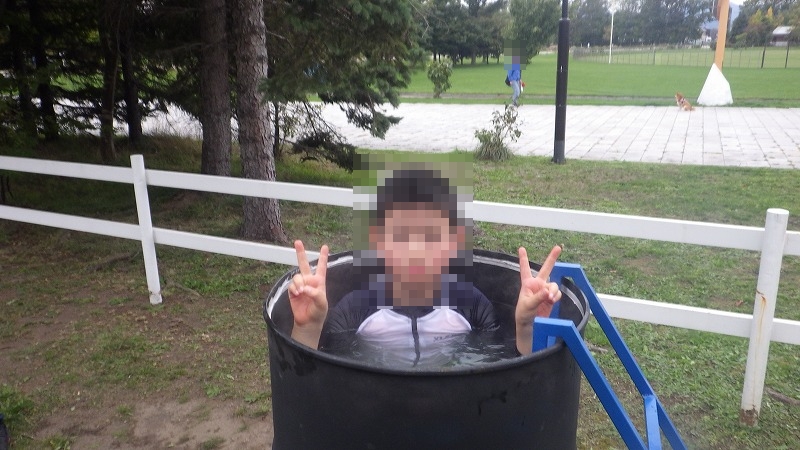 ドラム缶風呂に入った子供がピースをしている様子