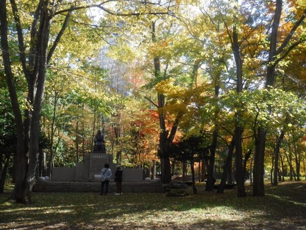 黄色に色づいた樹林地の中に佇む木下成太郎像と、鑑賞する人