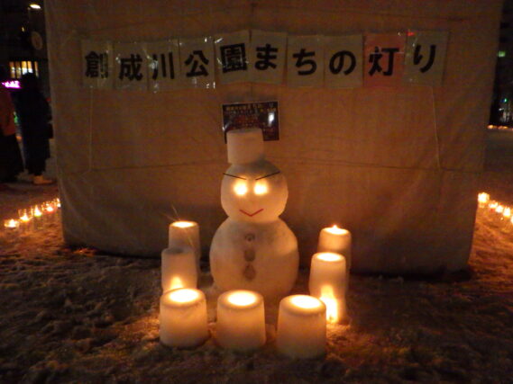 雪だるまの周りでスノーキャンドルが点灯されている様子