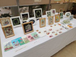 壁面に押し花で風景や鳥などを作った額の作品が並び、手前の机には小さな額の作品と葉が切作品が並ぶ。