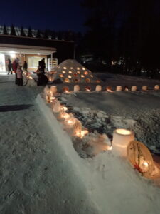 雪の低い棚の上に花の入ったアイスボードと雪で作ったスノーキャンドルが置かれ、ろうそくの明かりがともっている。奥の方に三角錐の山があり、そこの傾斜にもろうそくの明かりが見える。